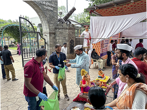 A group of people gathers outdoors for a Hindu festival, with several small Ganesha idols displayed. A man in traditional clothing places an offering into a green bag held by another person. In the background, a large stone archway with a cannon on top is visible.