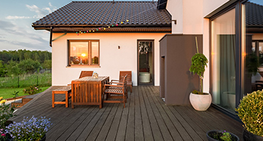 Back patio of a modern house with a dark wooden deck, a wooden table and chairs, and sunset light reflecting in a window above the deck.