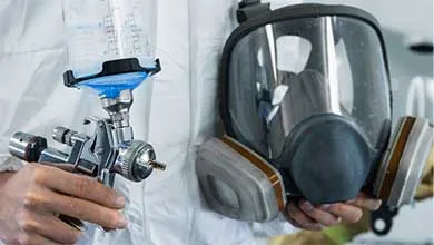 Person in white protective suit holding a paint spray gun and a full-face respirator mask.