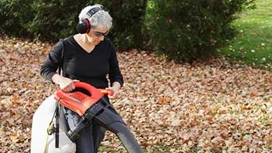 Woman with short gray hair, ear protection, and sunglasses, uses a red and black leaf blower to clear autumn leaves from a yard.