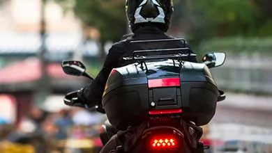 Rear view of a motorcyclist in a helmet and black jacket, riding a motorcycle with a large black storage case and illuminated red taillight.