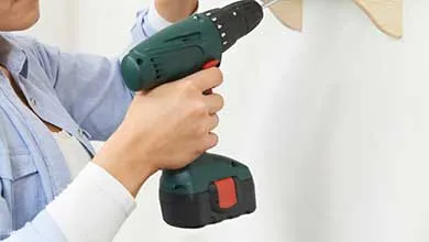 A person uses a cordless drill to attach a wooden bracket to a white wall.