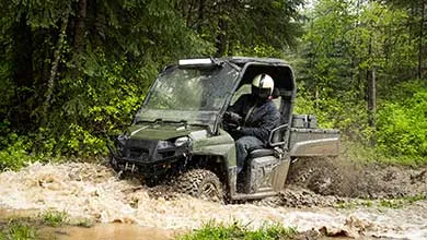 A helmeted person drives a green utility vehicle through a muddy forest puddle, splashing water and mud.