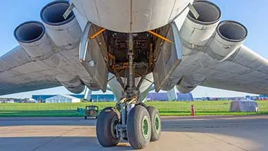 Low-angle close-up of a large airplane from below, showing its front landing gear and multiple jet engines under the wings on a sunny day.