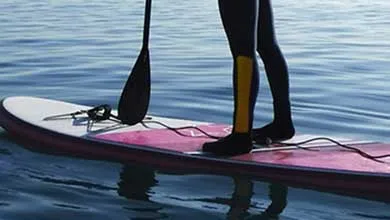 A person in a black wetsuit with a yellow stripe on one leg stands on a white and pink paddleboard floating on calm blue water. A black paddle rests upright beside their legs, and a coiled leash is attached near their feet.