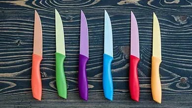 Six kitchen knives with orange, light green, purple, blue, red, and yellow handles arranged vertically on a dark wooden surface.