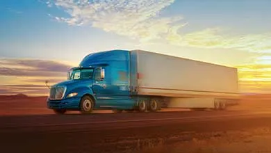 A blue semi-truck with a white trailer drives on an empty highway at sunset, with golden light on a dry landscape and scattered clouds in the sky.
