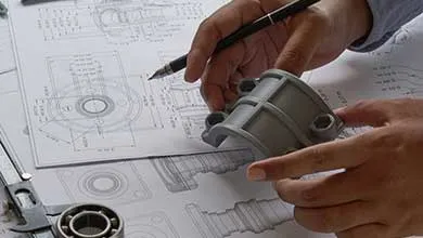 Hands hold a gray mechanical part with mounting holes over detailed engineering blueprints, while one hand holds a black pen above the drawings. A metal caliper is partially visible in the workspace's bottom corner.