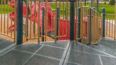 A dark green perforated metal playground platform with beige and green railings, and a red slide in the background.
