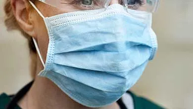 Close-up of a person wearing a light blue surgical mask, clear safety goggles, and green medical scrubs.