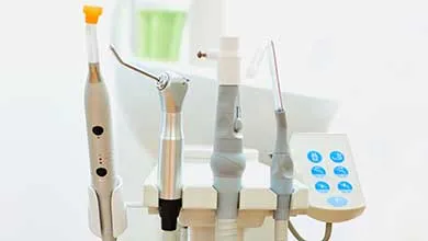 Dental equipment setup with a curing light (orange tip), handpiece, ultrasonic scaler, and probe on a white stand, next to a control panel with blue circular buttons on the right.