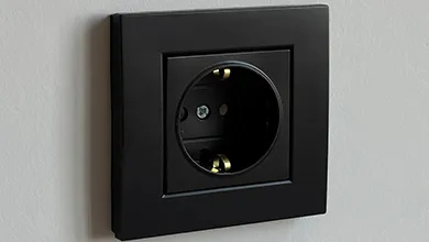 Matte black square electrical outlet with brass grounding clips on a light-colored wall.